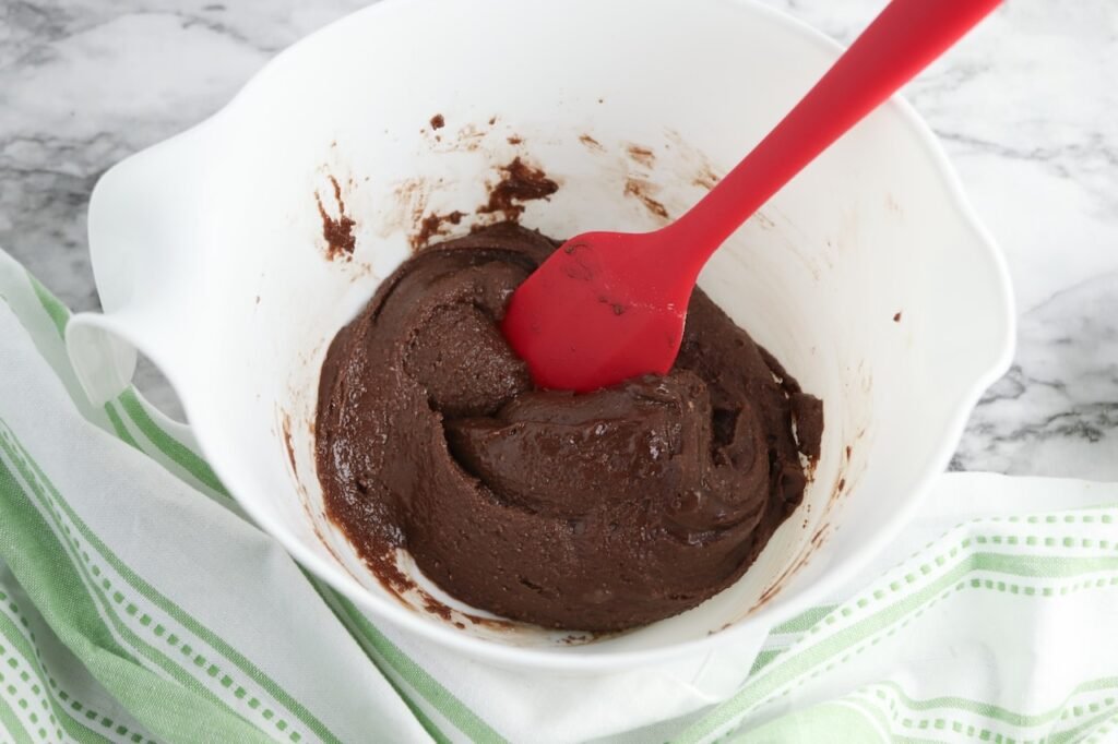 A thick brownie batter mixture in a white mixing bowl with a red silicone spatula, prepared by combining brownie mix, egg, and vegetable oil for brownie cinnamon rolls. The rich chocolate batter has a smooth, fudgy consistency and is ready to be spread over cinnamon roll dough for baking.