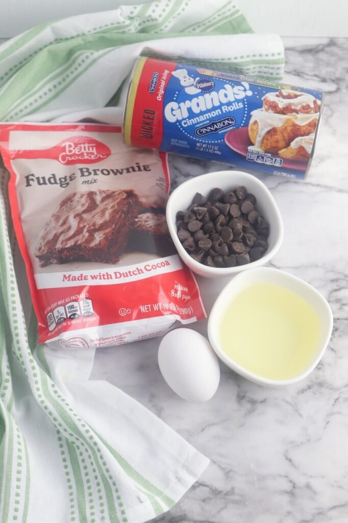 A flat lay of the ingredients needed to make brownie cinnamon rolls, including a package of Pillsbury Grands cinnamon rolls, a bag of fudge brownie mix, a bowl of chocolate chips, vegetable oil, and one egg arranged on a white marble surface with a green-striped kitchen towel in the background. This ingredient photo shows everything needed for this easy 5-ingredient brownie cinnamon roll recipe before mixing and baking.