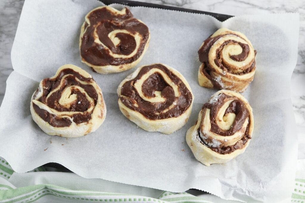 Unbaked dessert arranged on a parchment-lined baking sheet, showing thick swirls of chocolate filling layered inside the roll dough before baking. The rolls are spaced apart and ready to go into the oven for baking.