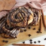Finished brownie cinnamon rolls stacked on a wooden board and topped with melted icing and a drizzle of chocolate. The baked rolls have thick chocolate brownie swirls inside and are surrounded by chocolate chips and cinnamon sticks, showing the gooey texture and rich chocolate layers.