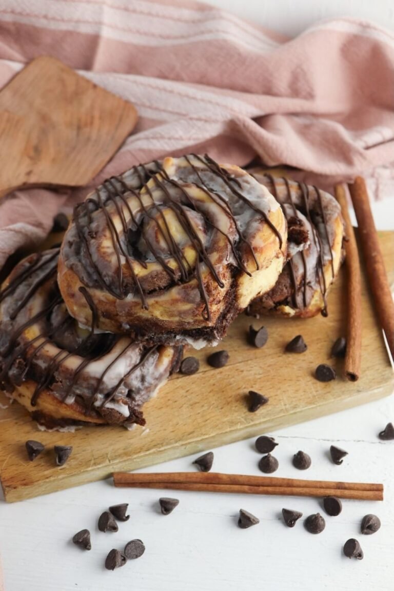 Finished brownie cinnamon rolls stacked on a wooden board and topped with melted icing and a drizzle of chocolate. The baked rolls have thick chocolate brownie swirls inside and are surrounded by chocolate chips and cinnamon sticks, showing the gooey texture and rich chocolate layers.