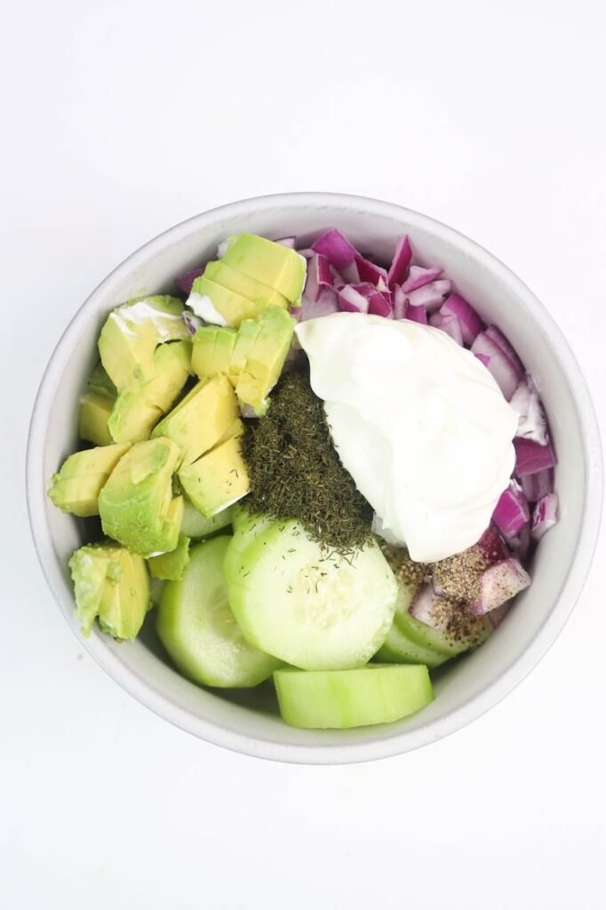 Overhead photo of sliced cucumbers, cubed avocado, diced red onion, sour cream, dill, salt, and black pepper in a white bowl before mixing.