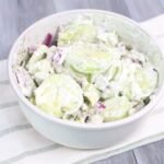A close-up side view of creamy avocado cucumber salad in a white bowl, made with thick cucumber slices, cubed avocado, diced red onion, and fresh dill mixed in a creamy sour cream dressing, served chilled on a light cloth over a wooden table.