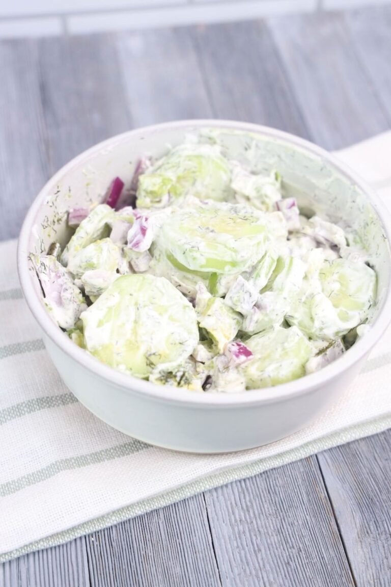 A close-up side view of creamy avocado cucumber salad in a white bowl, made with thick cucumber slices, cubed avocado, diced red onion, and fresh dill mixed in a creamy sour cream dressing, served chilled on a light cloth over a wooden table.