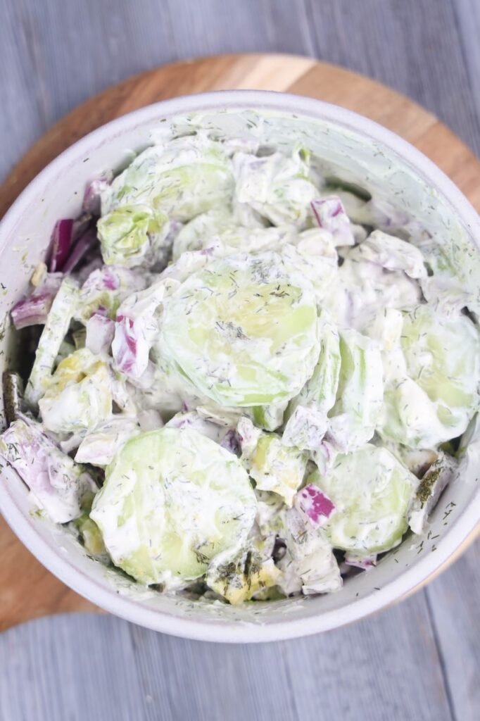 A close-up overhead view of creamy avocado cucumber salad in a white bowl, featuring thick slices of peeled cucumber, chunks of avocado, diced red onion, and fresh dill coated in a creamy sour cream dressing. The bowl sits on a wooden board over a gray wooden table, showing the fresh texture and creamy consistency of the chilled salad.