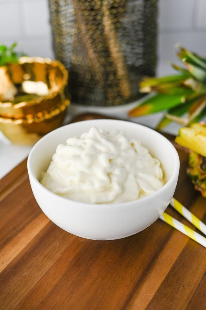 Close-up of homemade Dole Whip served in a white bowl on a wooden board, featuring a creamy soft-serve swirl with a smooth texture. Fresh pineapple pieces and striped paper straws in the background highlight the tropical flavors of this easy Disney-inspired frozen dessert.