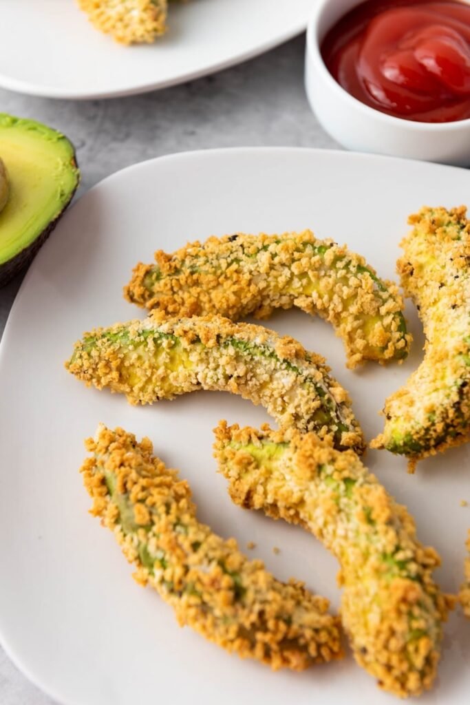 Close-up of crispy Air Fryer Avocado Fries on a white plate with a side of dipping sauce. The avocado wedges are coated in a golden breadcrumb crust that turns crunchy in the air fryer while the avocado inside stays creamy and tender.