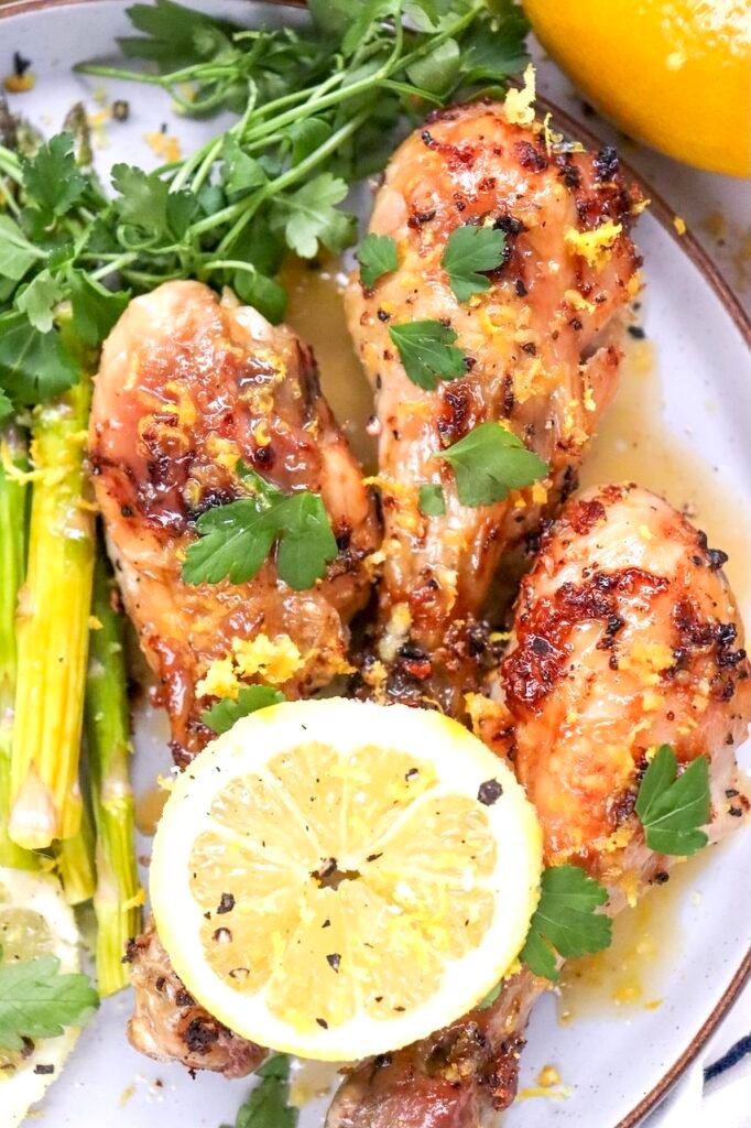 Close-up view of crispy lemon herb air fryer chicken legs garnished with fresh parsley, lemon zest, and a lemon slice. The chicken has golden-brown skin and is served with asparagus on a white plate with a light lemon sauce. This image highlights the texture and finished appearance of the cooked chicken drumsticks.