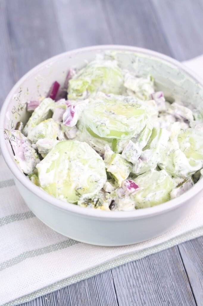 A side-angle view of creamy avocado cucumber salad in a white bowl, featuring thick slices of peeled cucumber, diced red onion, avocado chunks, and fresh dill coated in a creamy sour cream dressing. The bowl is set on a white striped cloth over a gray wooden table, showing the smooth texture and fresh ingredients of the chilled salad.