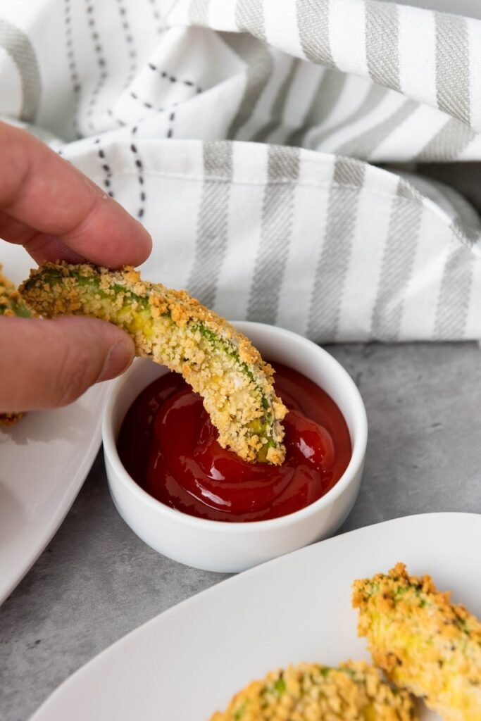 A hand dips a crispy Air Fryer Avocado Fry into a small bowl of dipping sauce. The avocado wedge is coated in a golden breadcrumb crust with a creamy green avocado center visible beneath the crunchy coating, showing the finished texture and serving suggestion.