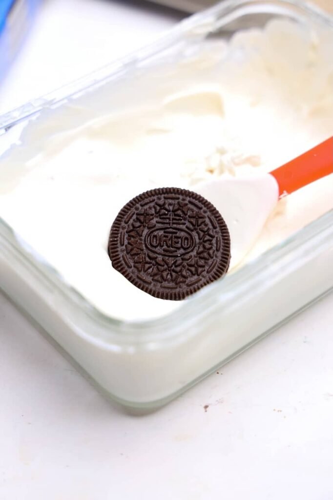 An Oreo cookie sits on a spatula above a dish of smooth melted white candy melts, demonstrating the process of coating the cookies for homemade Jack Skellington Oreos. This step helps create the white base needed for decorating the Halloween-themed Oreo treats.