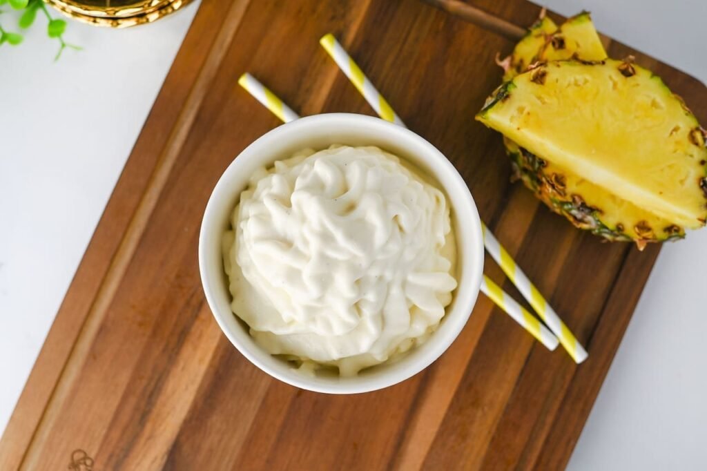 Overhead view of a bowl filled with homemade Dole Whip swirled into a soft-serve shape and served on a wooden board with fresh pineapple and striped paper straws. The creamy pineapple dessert has a smooth texture and is ready to enjoy as a refreshing Disney-inspired frozen treat.