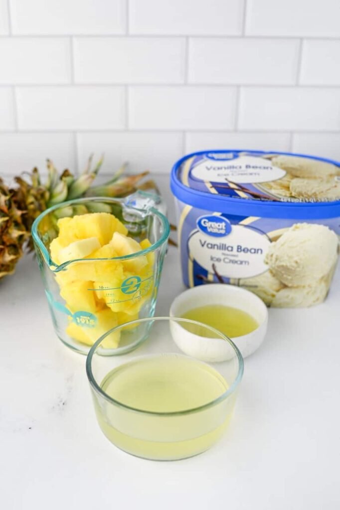 Ingredients for homemade soft serve arranged on a kitchen counter, including frozen pineapple chunks, pineapple juice, vanilla bean ice cream, and lemon juice before blending into a creamy pineapple soft serve dessert.