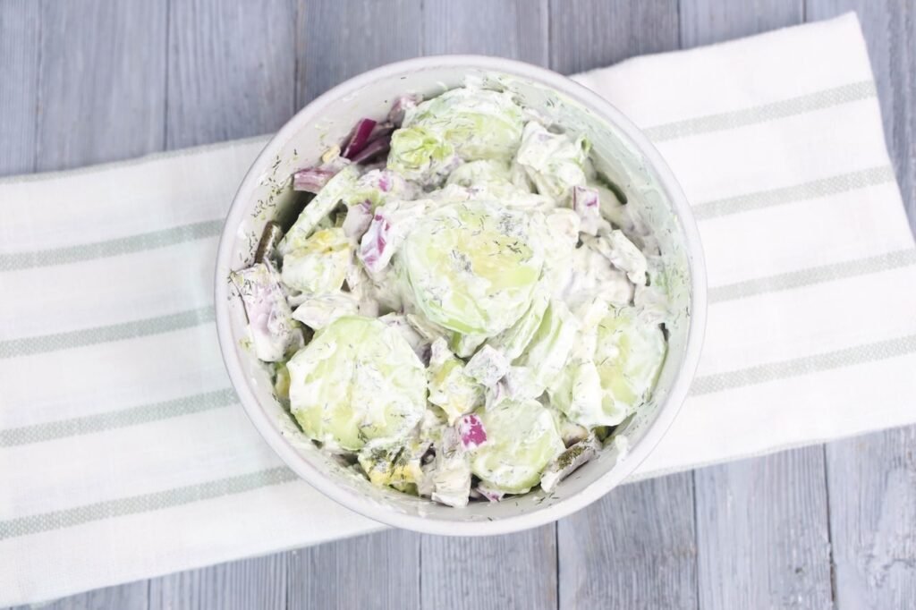 An overhead view of side dish served in a white bowl, made with sliced peeled cucumbers, diced red onion, avocado chunks, and fresh dill mixed in a creamy sour cream dressing. The bowl is placed on a white striped kitchen towel over a gray wooden table, showing the full plated salad ready to serve cold.