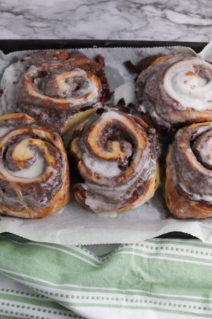 Freshly baked brownie cinnamon rolls on a parchment-lined baking sheet, topped with melted icing while still warm. The rolls show swirls of soft cinnamon roll dough layered with rich brownie filling, creating a gooey chocolate center and golden baked edges.