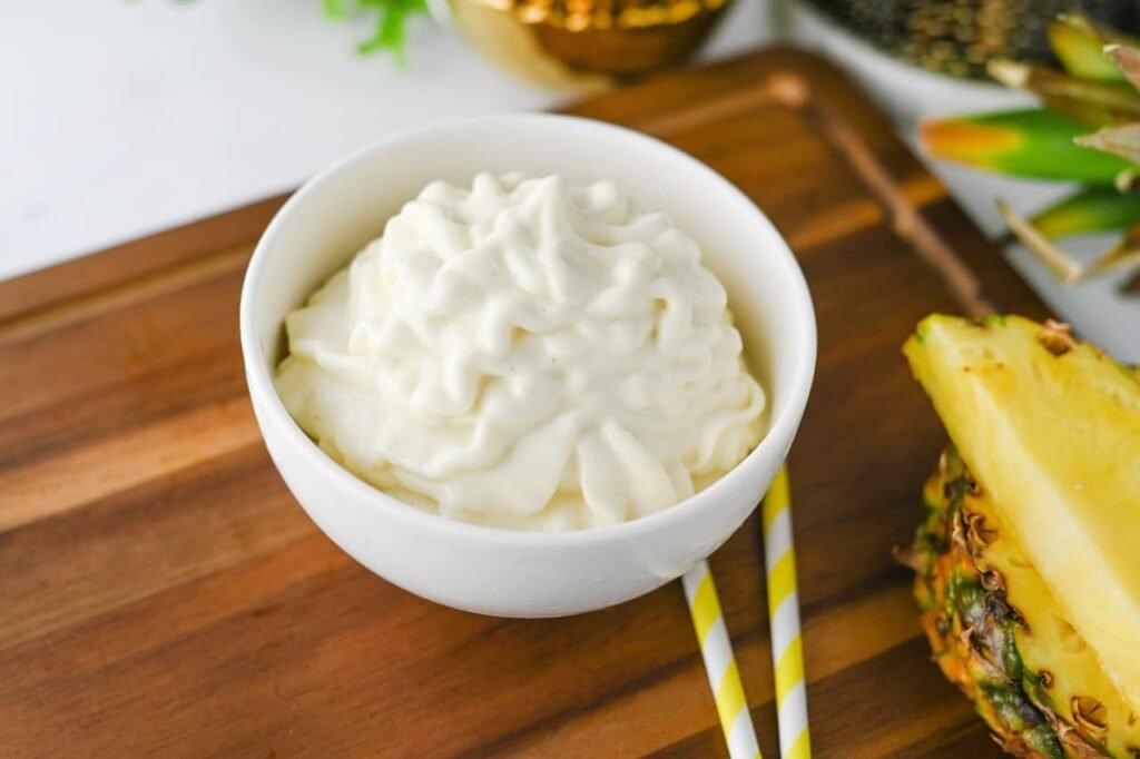 A bowl of homemade Dole Whip with a creamy soft-serve swirl sits on a wooden serving board next to fresh pineapple slices and striped paper straws. This Disney-inspired pineapple dessert is smooth, refreshing, and ready to serve immediately after blending.