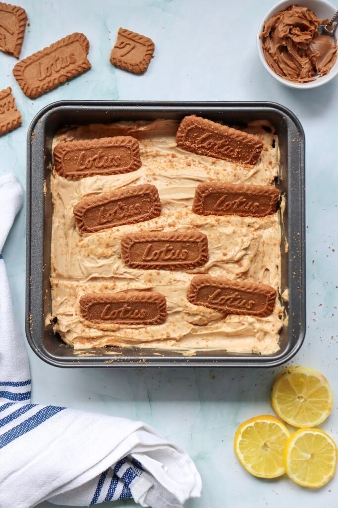 Overhead view of a square pan filled with no-bake Biscoff cheesecake topped with whole Lotus Biscoff cookies and a light sprinkle of cookie crumbs. The creamy cookie butter cheesecake filling has been smoothed into an even layer and decorated before chilling and serving.