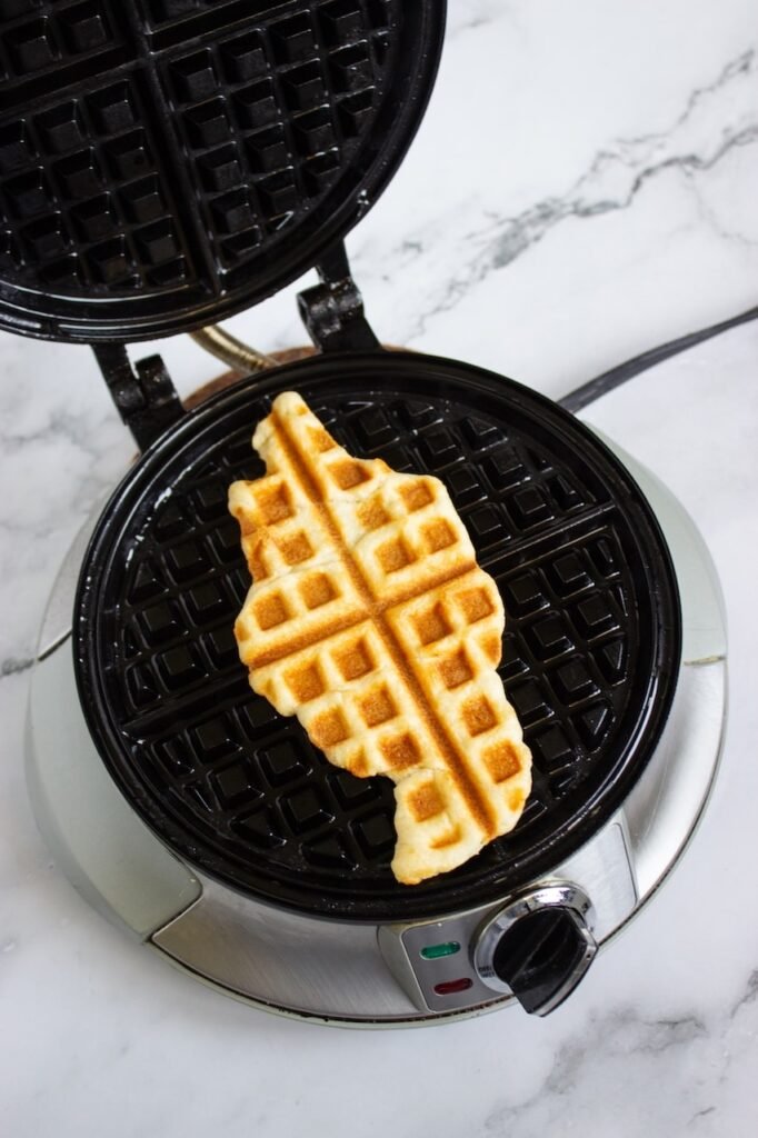 A golden brown croffle cooking inside an open waffle iron on a marble countertop, showing the crescent roll dough pressed into crisp waffle-like layers. This step creates the croffle’s crispy outside and soft, flaky inside before adding Biscoff whipped cream, strawberries, and cookie toppings.