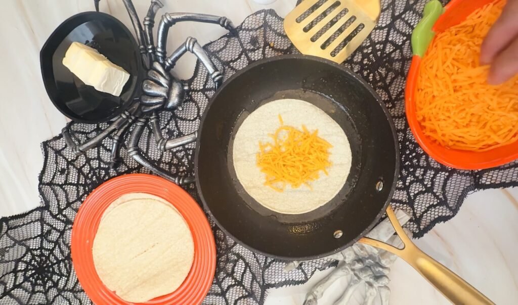 Shredded cheddar cheese is added to a tortilla in a skillet while making easy diiner, with Halloween decorations and additional ingredients arranged nearby.