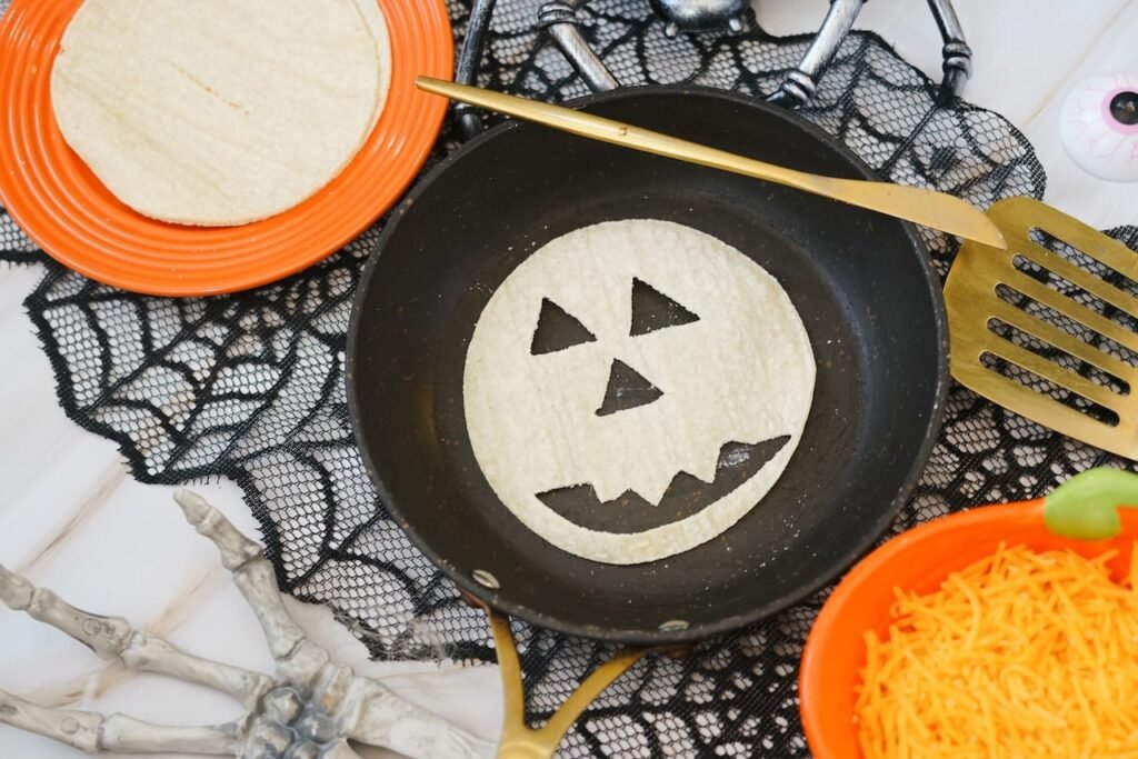 A tortilla carved into a face sits in a skillet during the preparation of Halloween quesadillas, surrounded by shredded cheddar cheese, extra tortillas, and festive Halloween decorations.