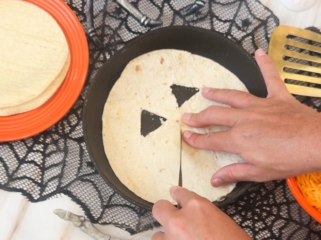 Hands place a tortilla with a carved jack-o-lantern face onto a cheese-filled quesadilla in a skillet during the assembly process for Halloween-themed quesadillas.