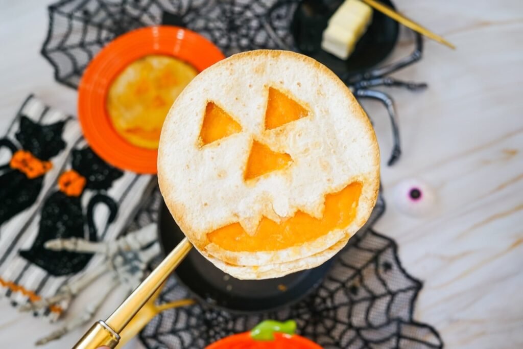 A finished dinner with melted cheddar cheese showing through a carved pumpkin face is held above a skillet with Halloween decorations in the background.