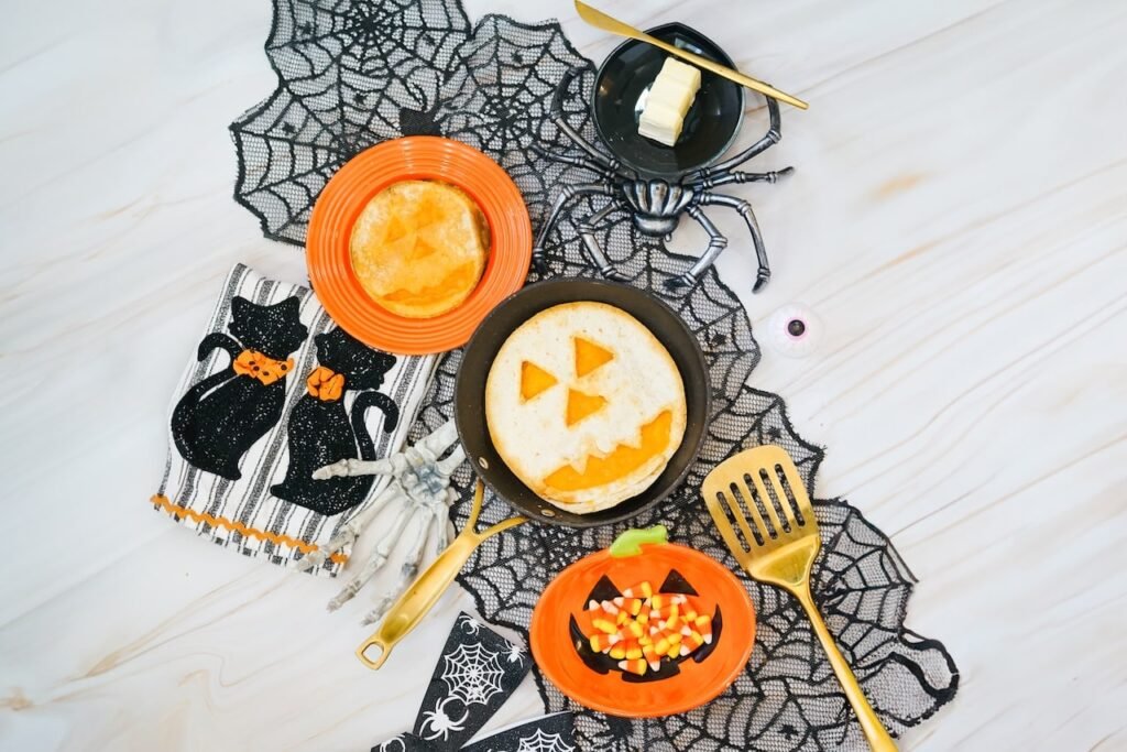 Overhead view of finished Jack-O-Lantern Quesadillas displayed in a skillet and on a plate with Halloween decorations, candy corn, spiderweb fabric, and festive serving dishes arranged on a tabletop.