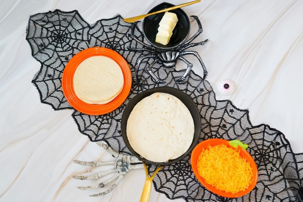 Ingredients for Jack-O-Lantern Quesadillas arranged on a Halloween-themed surface, including flour tortillas, shredded cheddar cheese, and butter beside a skillet and festive decorations before assembling the quesadillas.