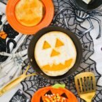 Overhead view of finished Jack-O-Lantern Quesadillas made with melted cheddar cheese and tortillas, displayed in a skillet with Halloween decorations including spiderweb fabric, candy corn, and festive serving dishes.