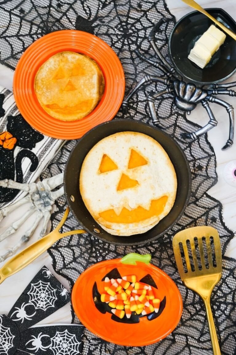Overhead view of finished Jack-O-Lantern Quesadillas made with melted cheddar cheese and tortillas, displayed in a skillet with Halloween decorations including spiderweb fabric, candy corn, and festive serving dishes.