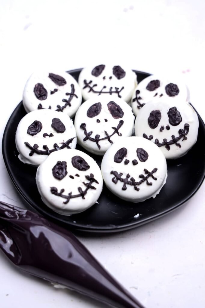 Finished Jack Skellington Oreos arranged on a black plate, each coated in white candy melts and decorated with black candy melts to create Jack Skellington's signature eyes, nose, and stitched smile. These easy no-bake Halloween treats are inspired by The Nightmare Before Christmas and are perfect for parties, dessert tables, and spooky movie nights.