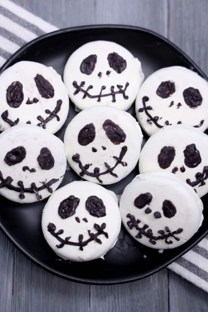 A black plate filled with homemade Jack Skellington Oreos decorated with white candy melts and black candy melt faces. Each Oreo features Jack Skellington's signature eyes, nose, and stitched smile, creating an easy Halloween dessert inspired by The Nightmare Before Christmas. The finished cookies are arranged on a serving plate and ready for a Halloween party or spooky movie night.