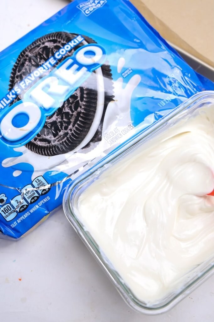 A package of Oreo cookies next to a glass dish filled with melted white candy melts being stirred with a spatula during preparation for halloween dessert.