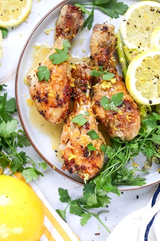Close-up of golden-brown air fryer chicken legs topped with fresh parsley and lemon zest on a serving platter with asparagus and sliced lemons. The chicken is coated in a light lemon garlic sauce and cooked until crisp on the outside while remaining juicy inside. This image highlights the finished chicken drumsticks ready to serve.