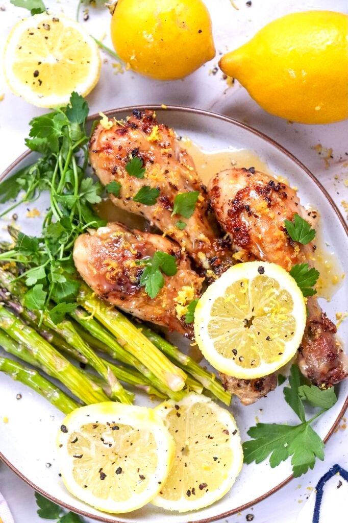 Close-up of crispy lemon herb air fryer chicken legs garnished with fresh parsley and lemon zest on a serving platter with asparagus and sliced lemons. The chicken has golden-brown skin and is drizzled with a light lemon sauce. This image showcases the finished air fryer chicken drumsticks ready to serve.