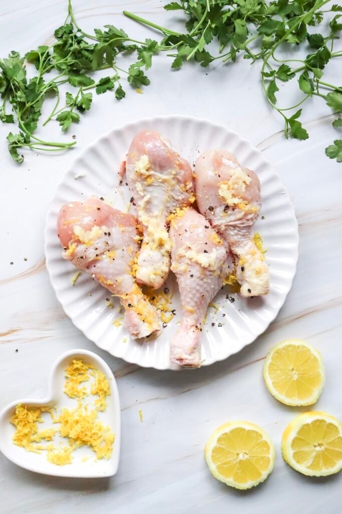 Raw drumsticks coated with fresh garlic, lemon zest, salt, and pepper on a white plate, surrounded by parsley and sliced lemons. This image shows the drumsticks after being seasoned and marinated before cooking in the air fryer.