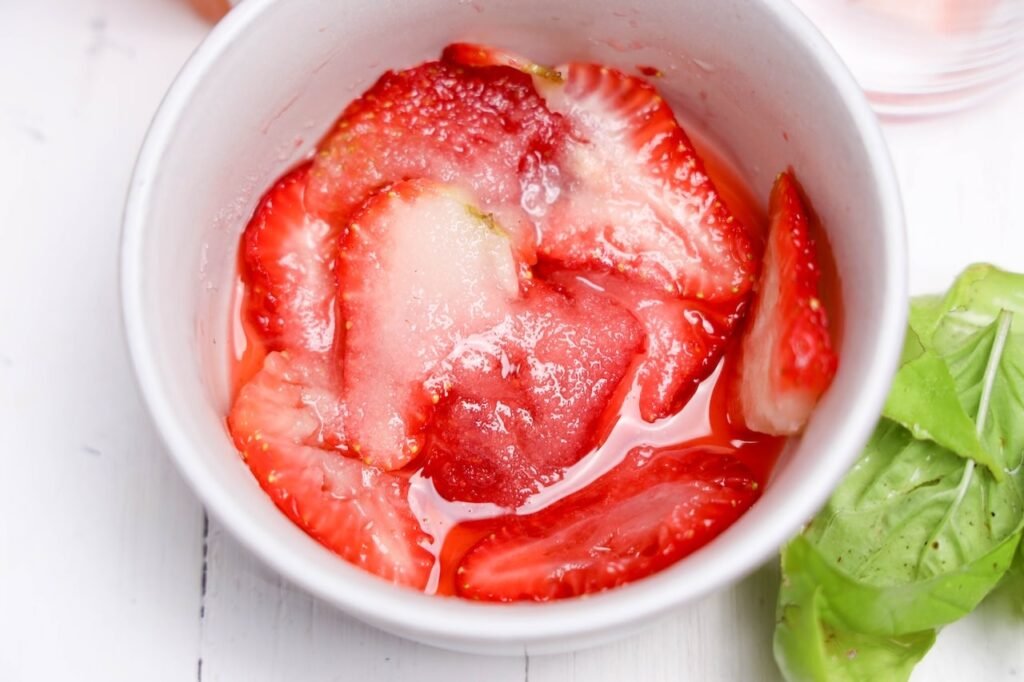 Sliced strawberries coated in sugar inside a small bowl with fresh basil leaves beside them for homemade strawberry basil lemonade preparation.