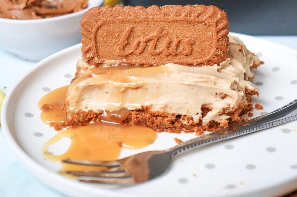 Close-up of a slice of no-bake Biscoff cheesecake served on a white plate with a buttery Biscoff cookie crust, creamy cookie butter cheesecake filling, a whole Lotus Biscoff cookie on top, and a drizzle of melted Biscoff spread. A fork rests beside the cheesecake slice, highlighting its rich, smooth texture.