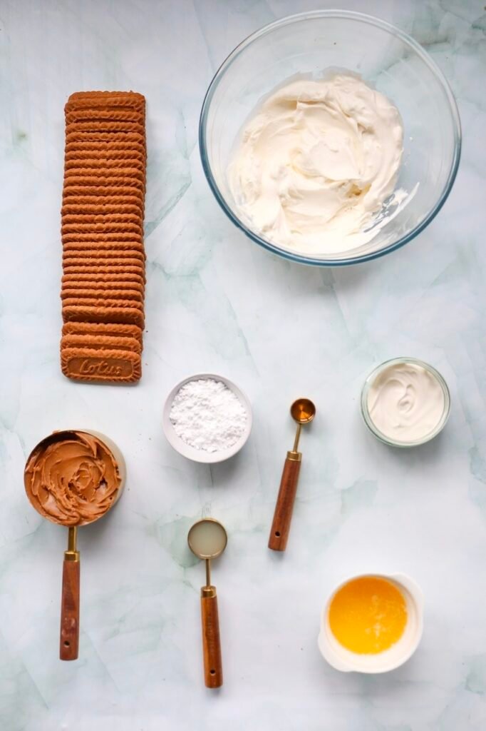 Overhead view of the ingredients for a no-bake dessert, including Lotus Biscoff cookies, Biscoff spread, cream cheese, whipping cream, powdered sugar, vanilla extract, and melted butter arranged on a light marble surface before preparation.