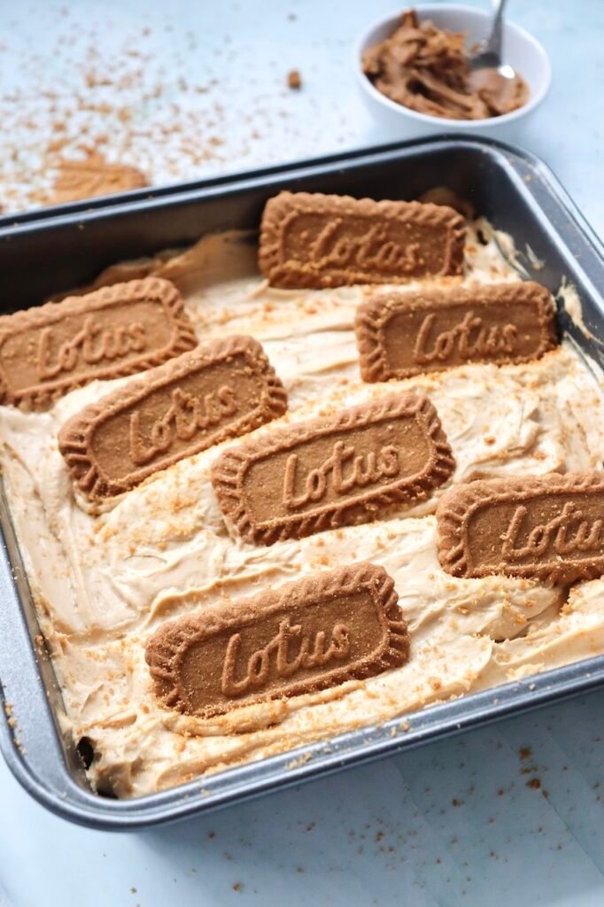 Close-up angled view of a square pan filled with no-bake Biscoff cheesecake. The creamy cookie butter cheesecake filling is smoothed into an even layer and topped with whole Lotus Biscoff cookies and crushed cookie crumbs before chilling. A small bowl of Biscoff spread is visible in the background.