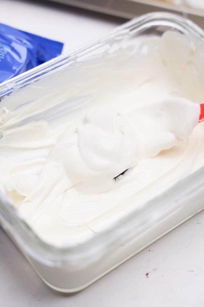 An Oreo cookie completely coated in melted white candy melts rests on a spatula above a glass dish of candy coating. This step in the recipe creates the smooth white base needed before piping on JS's signature eyes, nose, and stitched smile.
