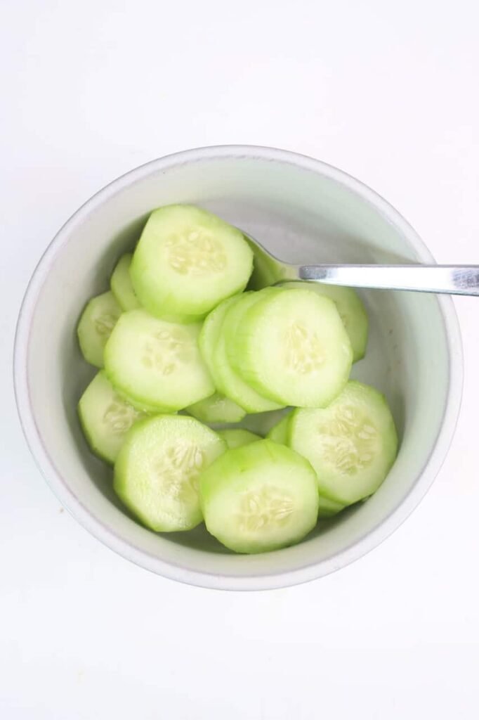 Close-up overhead photo of peeled cucumber slices in a white bowl with a spoon, prepared as the first step for making avocado cucumber salad.