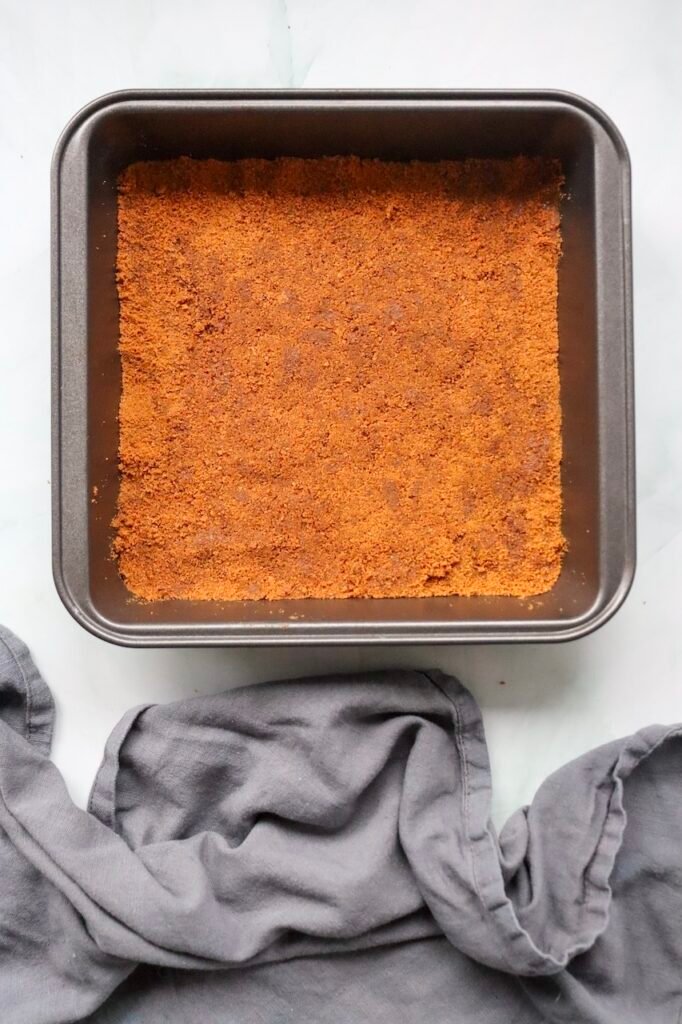 Overhead view of a square baking pan filled with a prepared Biscoff cookie crust made from finely crushed Lotus Biscoff cookies and butter. Bowls of powdered sugar and Biscoff spread sit nearby as the ingredients are assembled for a no-bake Biscoff cheesecake recipe.