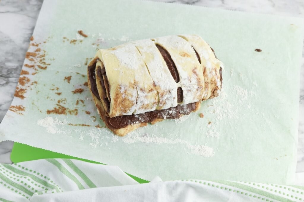 Cinnamon roll dough rolled up with brownie batter filling on floured parchment paper, showing the layered chocolate swirls before slicing into individual brownie cinnamon rolls. This step helps create the rich, fudgy center in each baked roll.