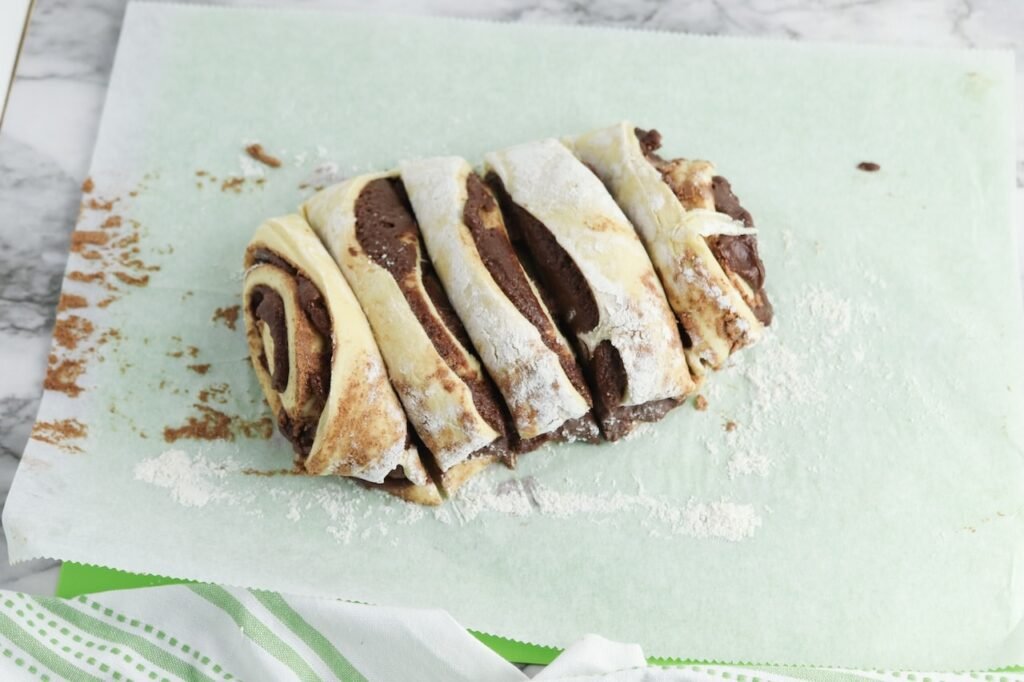 Sliced dough on floured parchment paper, showing thick layers of filling swirled inside the dough before baking. This step shows how the rolls are cut into individual pieces before being placed on the baking sheet.