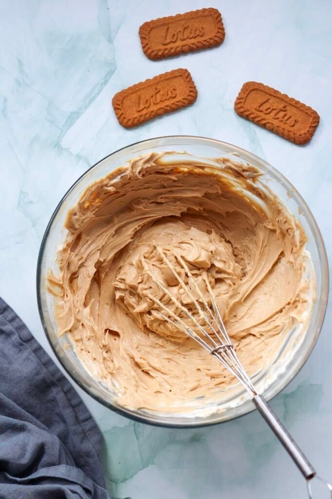 Overhead view of a smooth  filling in a glass mixing bowl after the cream cheese, spread, vanilla extract, and icing sugar have been mixed together. A whisk rests in the creamy cookie butter mixture, with Lotus cookies arranged nearby before whipped cream is folded into the filling.