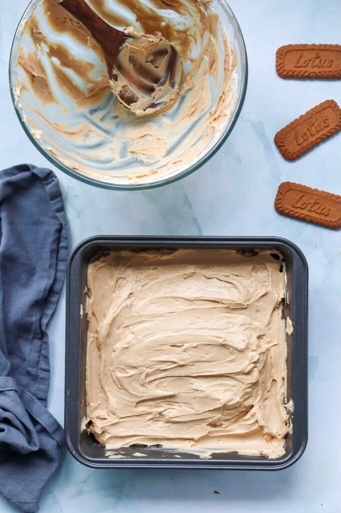 Overhead view of a square pan filled with smooth Biscoff cheesecake filling spread evenly over a Biscoff cookie crust. A mixing bowl with the remaining cookie butter filling and Lotus Biscoff cookies sits nearby as the no-bake cheesecake is prepared for chilling.