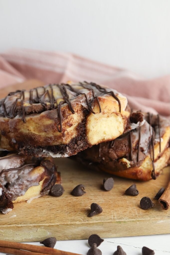 Side view of finished brownie cinnamon rolls stacked on a wooden board, drizzled with icing and melted chocolate. The image shows the thick fudgy brownie layer baked into the bottom and center of the soft cinnamon roll, highlighting the gooey chocolate filling and fluffy texture.