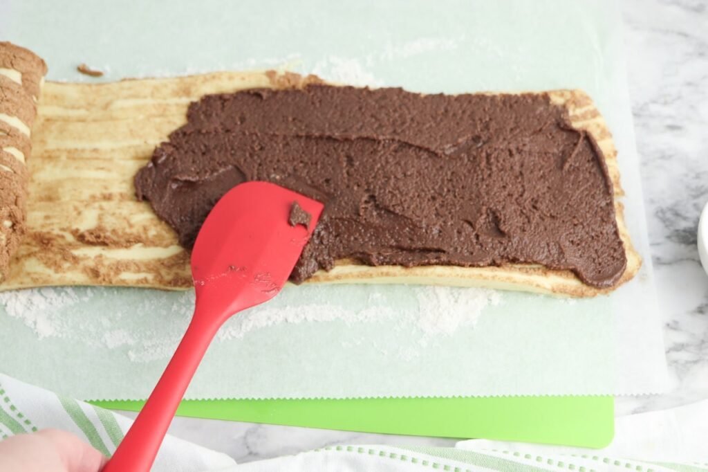 batter being spread over unrolled cinnamon roll dough with a red spatula on floured parchment paper. This step shows how to layer the thick chocolate filling evenly across the dough before rolling it up.