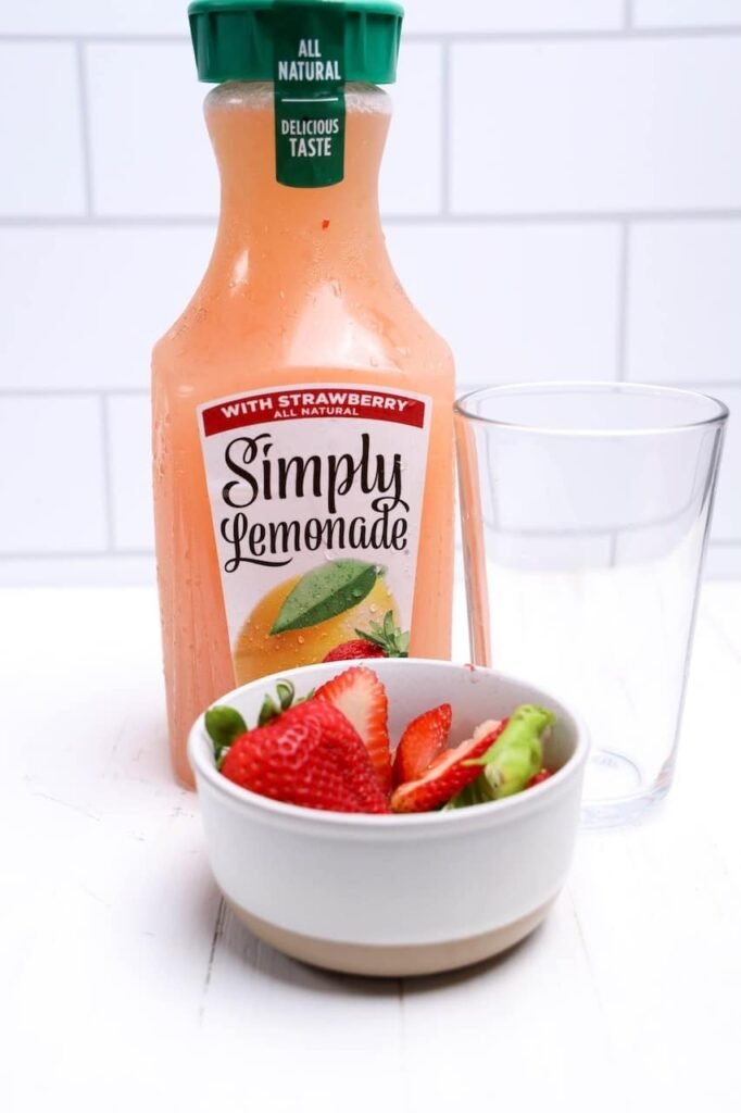 Bottle of Simply Strawberry Lemonade beside a glass and a bowl filled with fresh strawberries and basil leaves for homemade strawberry basil lemonade.